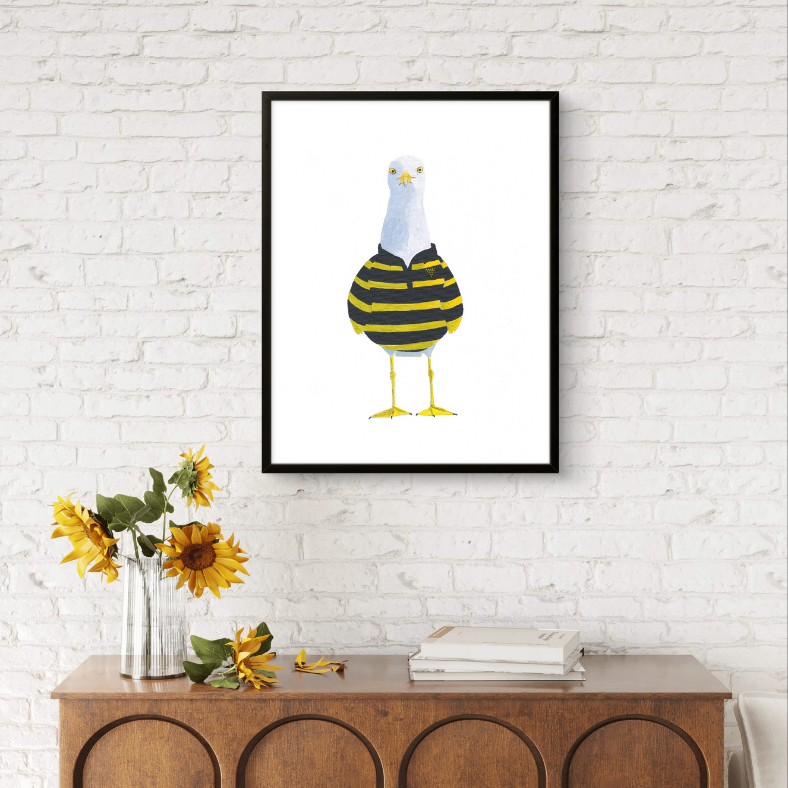Framed artwork of a cartoon seagull with a striped sweater on a white brick wall above a wooden console table with sunflowers and books.